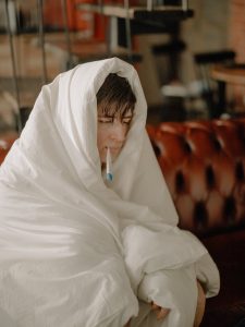 Ein junger Mann sitzt mit einer Decke und Fieberthermometer auf dem Sofa.
